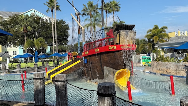 Parque acuático con área de juegos temática de barco pirata, que incluye toboganes y un puente colgante.