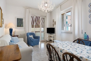 A room with a white sofa, a blue armchair, a wooden table, a TV on a stand, and a dining table with a patterned tablecloth. - Apartment 'Zaffiro  with Garage' with Private Terrace, Wi-Fi and Air Conditioning (Rapallo)