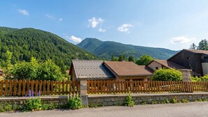 Ambiente rural com cerca de madeira, paredes de pedra e montanhas ao fundo.