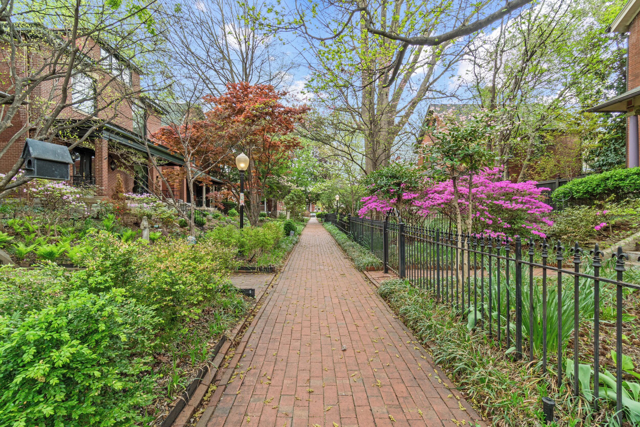 A brick pathway flanked by lush greenery and a black metal fence, leading towards a residential area with brick houses.