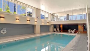Una piscina interior con agua azul cristalina, una zona soleada con sillones de descanso y vistas a los edificios exteriores a través de amplias ventanas.
