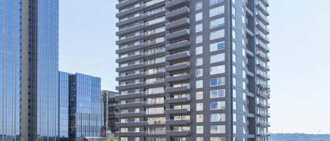 A modern high-rise building with multiple balconies, surrounded by other tall buildings and a parking lot with cars.