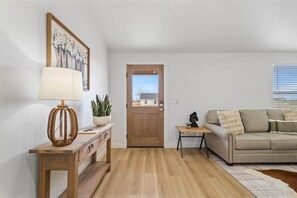 Un salon avec un plancher en bois, un canapé beige, une table basse, une lampe, une plante et une porte donnant sur l’extérieur.
