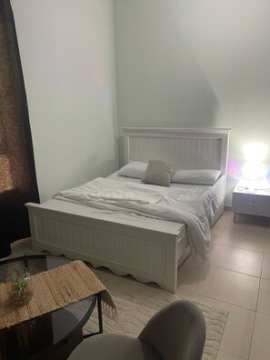 Ein Schlafzimmer mit einem weißen Bett, einem Nachttisch, einem Sessel und einem kleinen Tisch mit einer Pflanze.