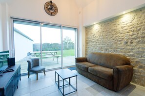 A living room with a stone wall, a brown leather sofa, a grey armchair, and a small black table. - Gîte du Cellier - Jardin et terrasse (Carentan)