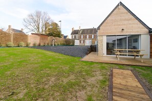 A modern house with a wooden deck, a large glass door, and a stone wall. - Gîte du Cellier - Jardin et terrasse (Carentan)