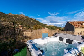 Un jacuzzi dans un jardin, entouré d’une clôture en bois, avec une montagne en arrière-plan et un bâtiment doté d’un toit en tuiles.