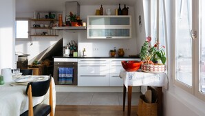 A modern kitchen with white cabinets, a dining area with a table and chairs, and a window with curtains. - Le Rooftop Aux Puces - Terrasse rooftop (Saint Ouen)