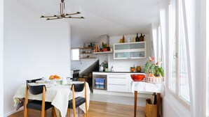 A modern kitchen with white cabinets, a dining area with a table set for two, and a view of the street through large windows. - Le Rooftop Aux Puces - Terrasse rooftop (Saint Ouen)
