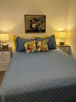 Un dormitorio con una cama grande, dos mesitas de noche con lámparas, un cuadro floral en la pared y una colcha acolchada.