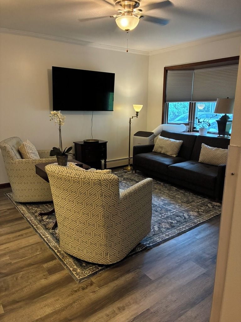 A living room with a flat-screen TV, two patterned armchairs, a dark sofa, a floor lamp, and a window with blinds.