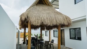 Een overdekt terras met rieten dak, een tafel en stoelen.