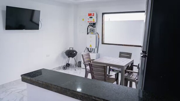 Een kleine kamer met een flatscreen televisie, een stopcontact, een elektrische installatie, een grill, een tafel met stoelen en een koelkast.