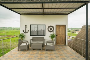 A covered outdoor seating area with a sofa and chairs, a wooden door, and a view of the countryside.