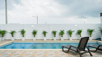 Una piscina con sillones de descanso, plantas en macetas y una pared blanca con líneas eléctricas.