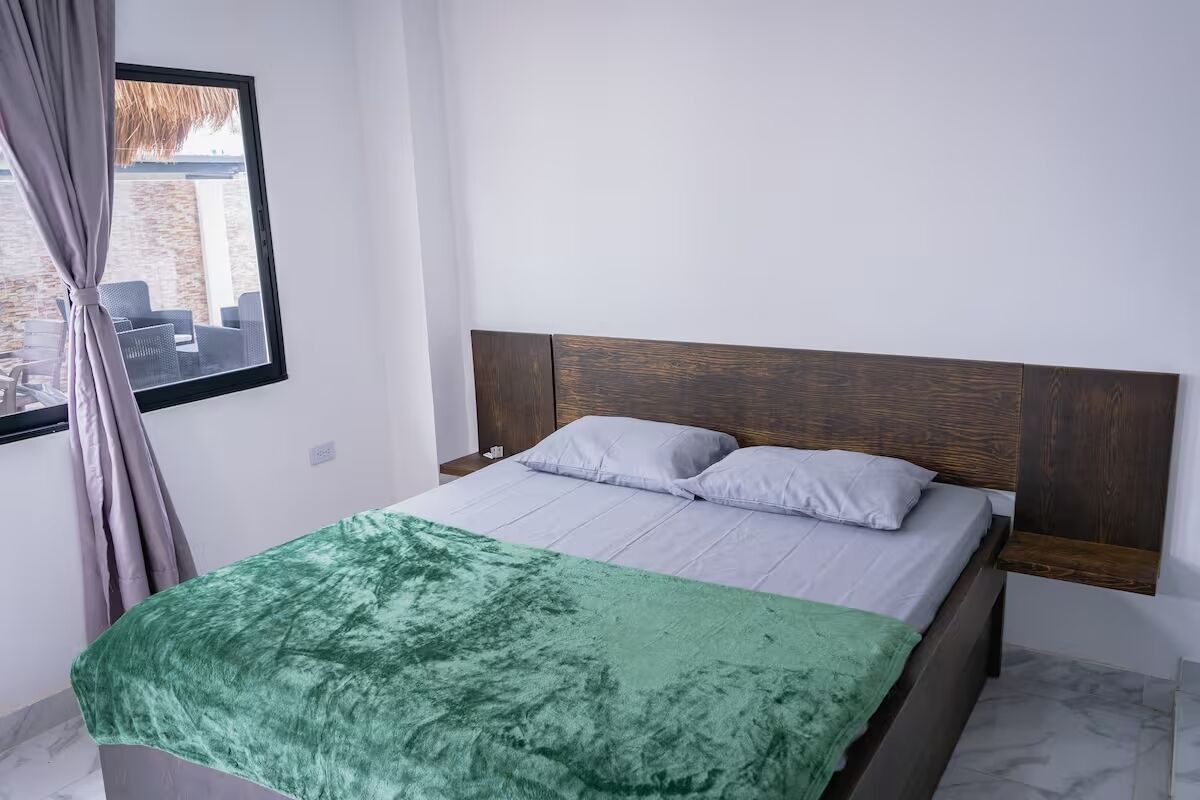 Un dormitorio con una cama, una manta verde, un cabecero de madera y una ventana con cortinas.