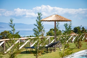 A poolside area with lounge chairs and a thatched umbrella, surrounded by greenery and overlooking a body of water. - Olive Grove Panorama Suite Pende (Kassandra)