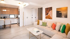A modern living room with a sofa, a coffee table, and a dining area with chairs. There is a kitchen visible in the background. - Olive Grove Panorama Suite Pende (Kassandra)