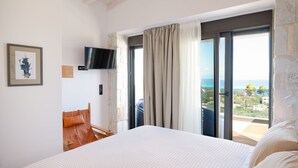 Een slaapkamer met een bed, een televisie aan een stenen muur, een stoel en uitzicht op zee door grote ramen.