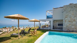 Een zwembadgedeelte met ligbedden en rieten parasols, een stenen muur en uitzicht op de zee.