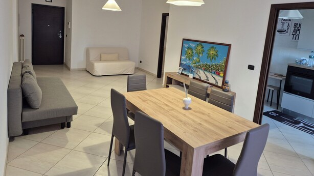 Ein modernes Wohnzimmer mit einer Couch, einem Esstisch und Stühlen. An der Wand hängt ein Bild und durch eine offene Tür ist eine Küchenzeile zu sehen.