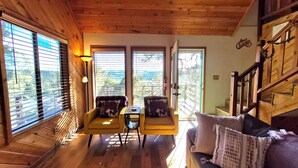Interior de cabana com piso de madeira, um sofá, duas poltronas, uma mesa e grandes janelas com persianas.