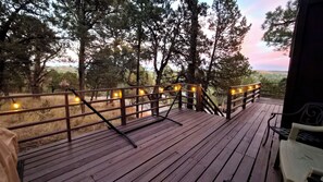 Une terrasse en bois avec une rambarde, des guirlandes lumineuses et une vue sur les arbres et les collines.