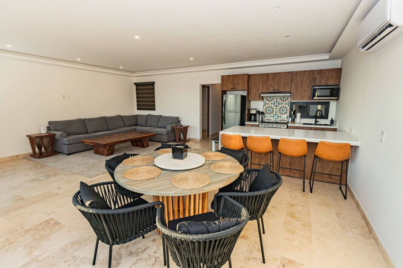 A modern living room with a gray sectional sofa, a wooden coffee table, and a round dining table with woven placemats and black chairs.