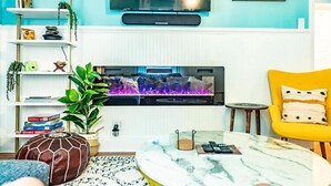 Olohuone, jossa on takka, takan yläpuolella seinälle asennettu televisio, keltainen nojatuoli ja hylly, jossa on erilaisia esineitä.