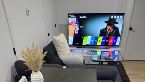 Moderni olohuone, jossa on harmaa puinen pöytä, valkoiset tuolit, lasinen sohvapöytä ja litteänäyttöinen televisio, jonka näytöllä näkyy Netflixin käyttöliittymä.