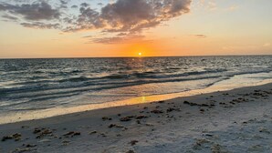 Ein Strand mit untergehender Sonne über dem Meer, sanfte Wellen, die am Ufer branden, und verstreutem Seetang im Sand.