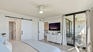 Ein Schlafzimmer mit Bett, Schminktisch, Nachttisch, Deckenventilator und einem großen Fenster mit Vorhängen.
