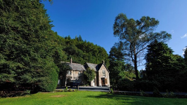 A stone building with a steep roof, surrounded by lush greenery and trees. - Ladye Park (Liskeard)