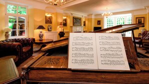 A grand piano with sheet music in a warmly lit room with chandeliers, leather furniture, and a fireplace. - Ladye Park (Liskeard)