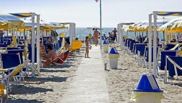 Een strand met rijen blauwe en gele strandstoelen en parasols, een heldere lucht en een vlaggenmast in de verte.