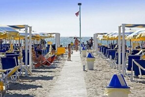 Ein Strand mit Reihen aus blauen und gelben Liegestühlen und Sonnenschirmen, einem klaren Himmel und einem Fahnenmast in der Ferne.