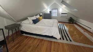 Ein Schlafzimmer mit schrägverdeckter Decke, einem Bett mit weißer Bettwäsche, einem kleinen Nachttisch, einem bei einem Bett stehenden Tisch, einem Fenster mit Jalousien, einer Klimaanlage und einer Topfpflanze.