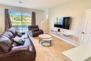 A living room with a leather sofa, a TV, a small table, and a view of a pool and greenery outside. - 4BR Pool Townhome at Bella Vida Resort (Kissimmee)