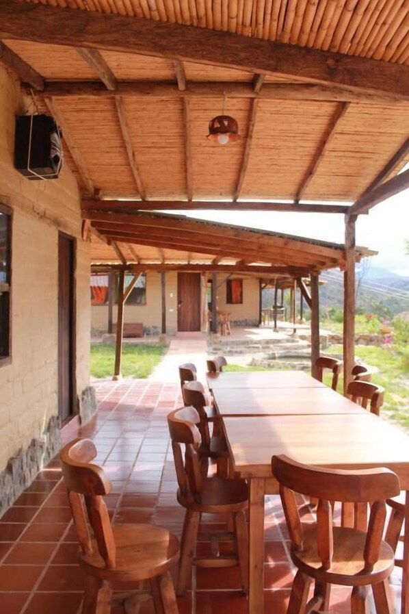 An outdoor dining area with wooden tables and chairs, a thatched roof, and a view of the surrounding landscape. - POSADA EL ARTESANO DE RAQUIRA (Ráquira)