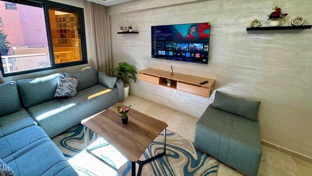 A modern living room with a sectional sofa, a coffee table, a TV mounted on the wall, and a chandelier. - Gueliz Twin Balcony Apartment F3 (Marrakesh)