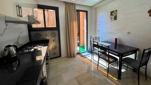 A compact kitchen with a gas stove, a kettle, and a window with curtains. There is a dining table with chairs and a red bottle on it. - Gueliz Twin Balcony Apartment F3 (Marrakesh)