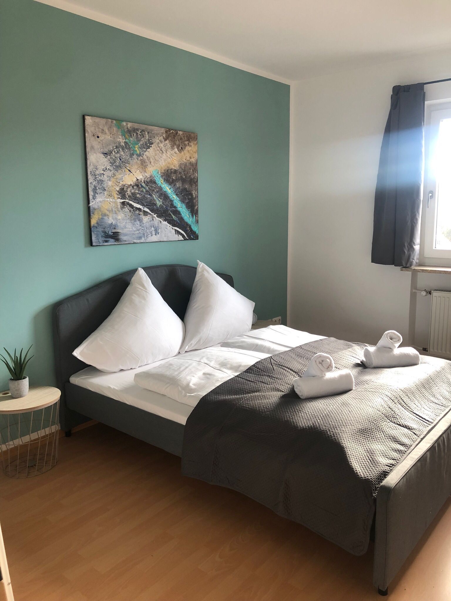 A neatly made bed with white linens and pillows, a gray headboard, and a textured blanket. There is a painting on the wall above the bed and a small potted plant on a side table.