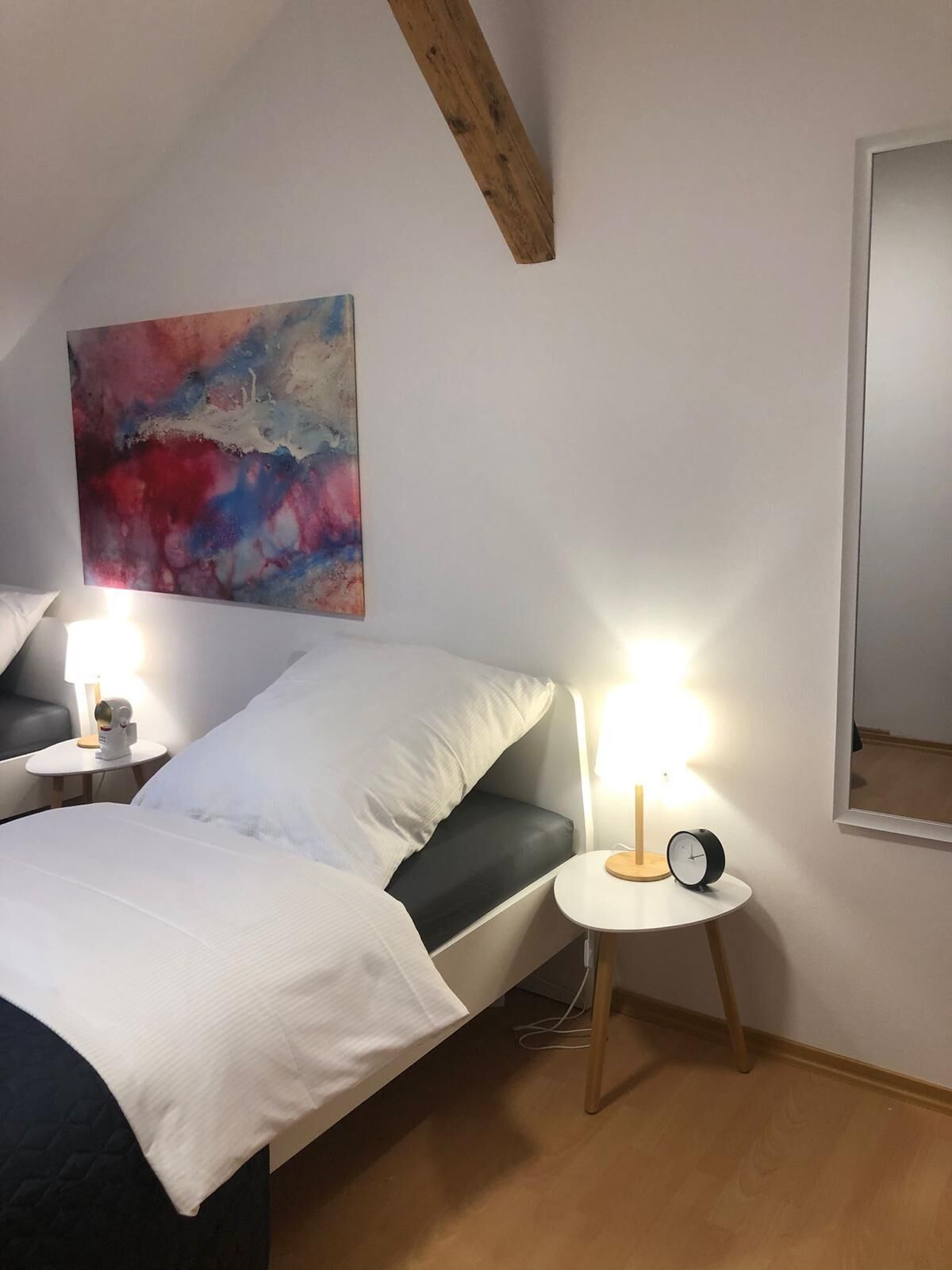 A bedroom with a bed, bedside lamps, a clock, a painting, and a wooden ceiling beam.