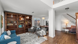 A spacious living room with a blue sofa, grey armchairs, a wooden bookshelf, and a staircase leading to an upper level. - Family Friendly Hurst Home with Salwater Poo,l Fireplace, and Spacious Layout (Hurst)