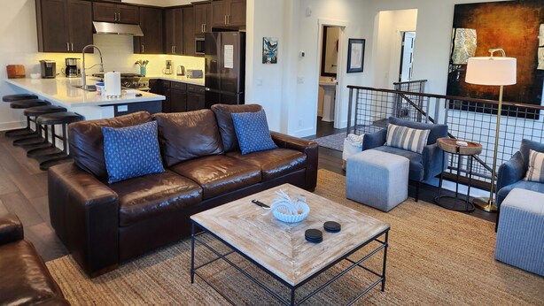 A modern living room with a brown leather sofa, a coffee table, and a dining area with dark wooden cabinets and a stainless steel refrigerator. - Snowcreek Resort Vacation Rental 1595 (Mammoth Lakes)