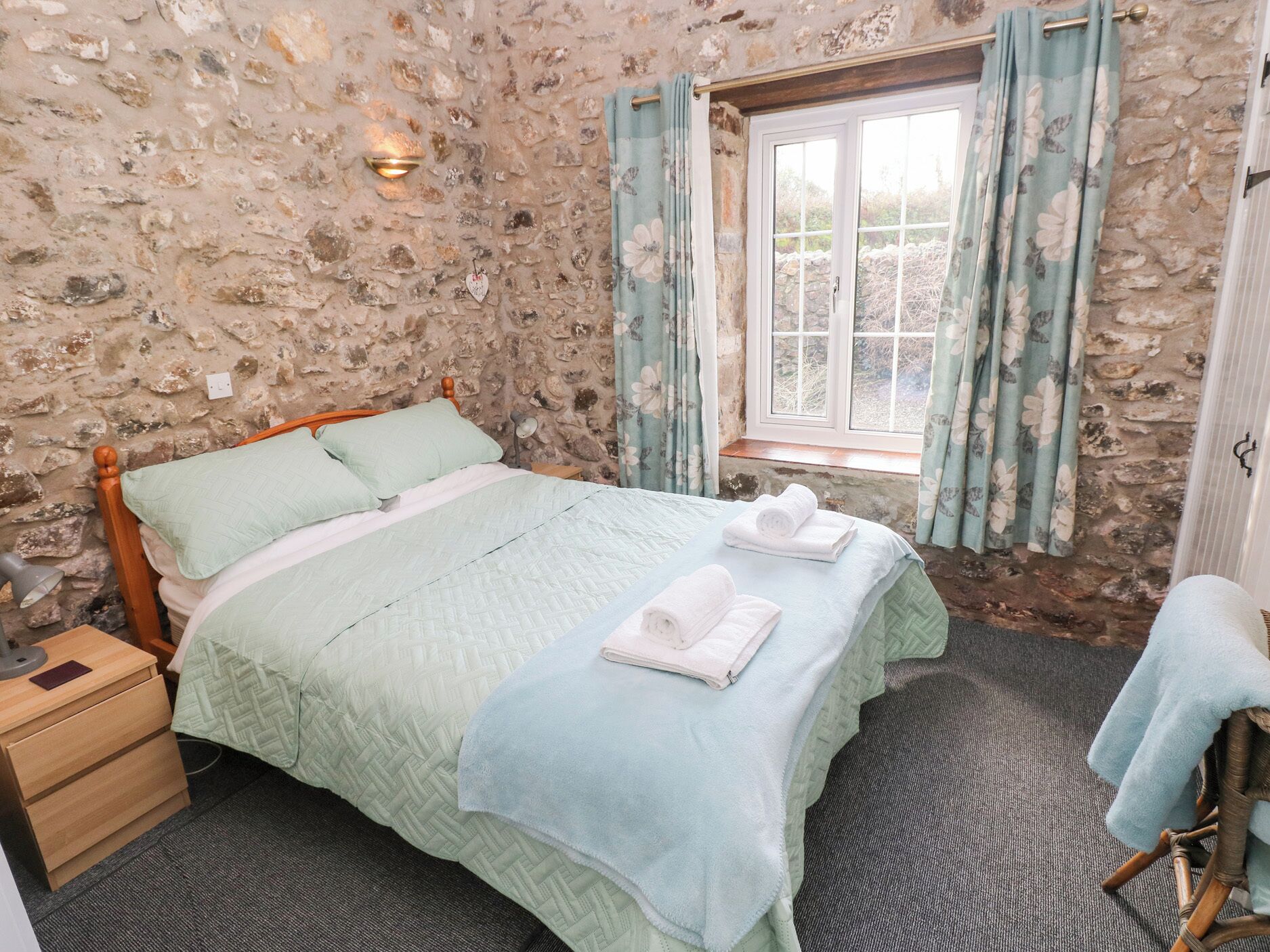 A bedroom with a stone wall, a wooden bed, a window with curtains, and a bedside table with a lamp.