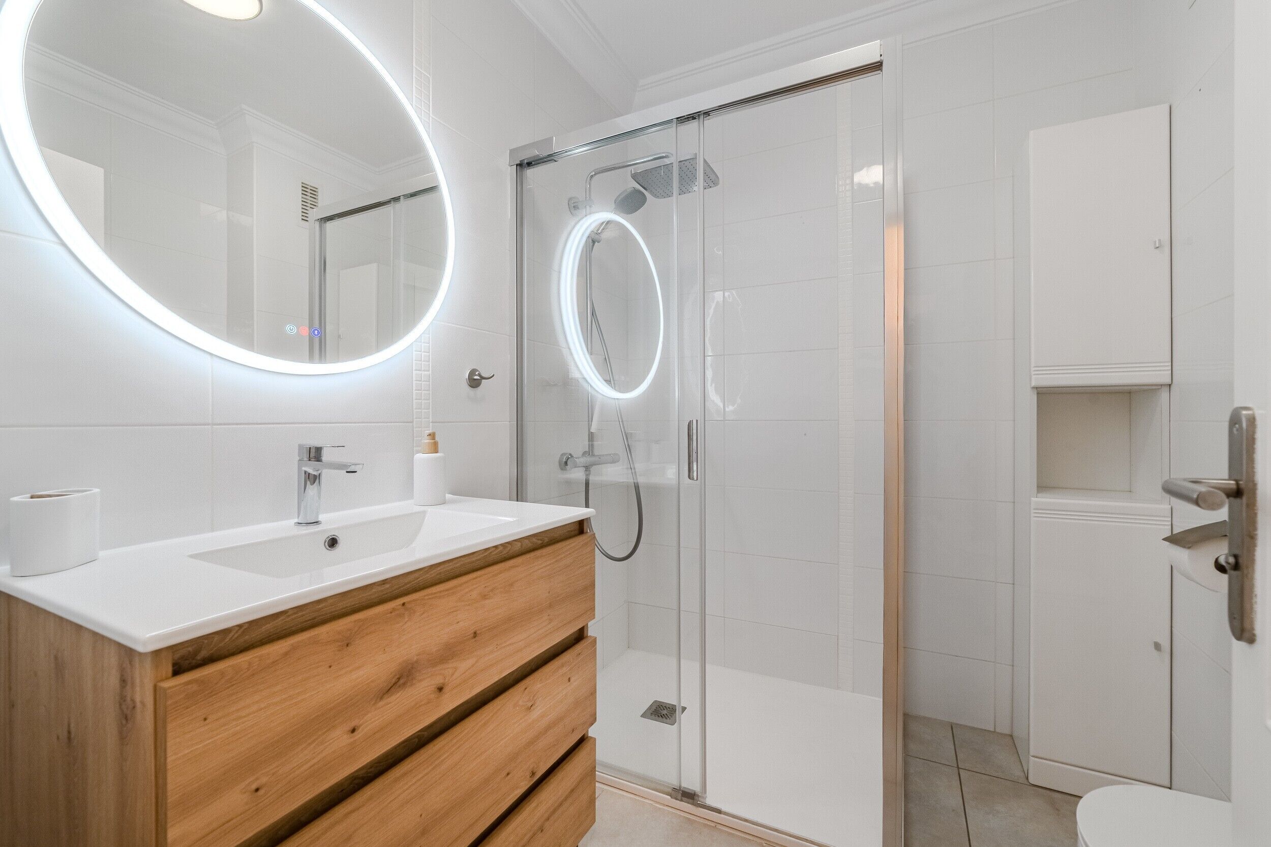 A modern bathroom with a glass-enclosed shower, a round mirror with lights, a wooden vanity, and a toilet.