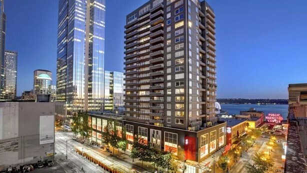 Een modern hoog gebouw met meerdere verdiepingen en balkons, omringd door andere hoge gebouwen en een waterkant.
