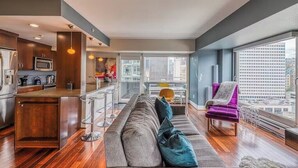Een modern appartement met keuken, eethoek en woonkamer. Het heeft een groot raam met uitzicht over de stad, een bank en een paarse fauteuil.