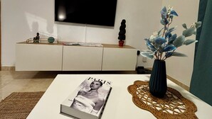 Un soggiorno moderno con una TV a schermo piatto montata a parete, un divano bianco, un tavolino da caffè con una rivista e una pianta decorativa.